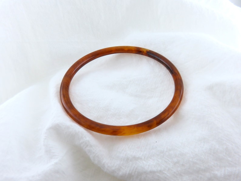 May include: A circular bangle bracelet featuring a translucent amber and brown tortoiseshell pattern. The bracelet has a smooth, polished finish and a rounded profile, showcasing a classic mottled design with varying shades of golden brown.