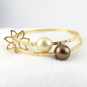May include: Gold-tone bangle bracelet featuring a floral design and two pearl accents. The bracelet has a hammered texture and includes a cream-colored and a brown pearl. The bracelet is a jewelry item.