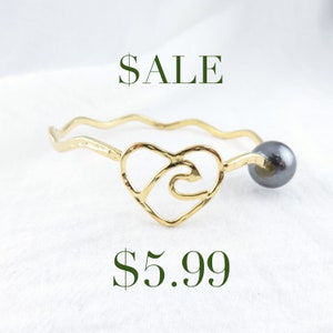 May include: Gold bracelet with a heart-shaped charm featuring a wave design and a black bead. The bracelet is on sale for $5.99.