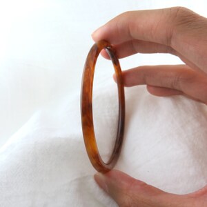 May include: A thin, circular bangle bracelet featuring a polished, translucent amber-colored resin material with a marbled, swirling pattern of dark brown and golden honey tones throughout the smooth, rounded surface of the jewelry piece.
