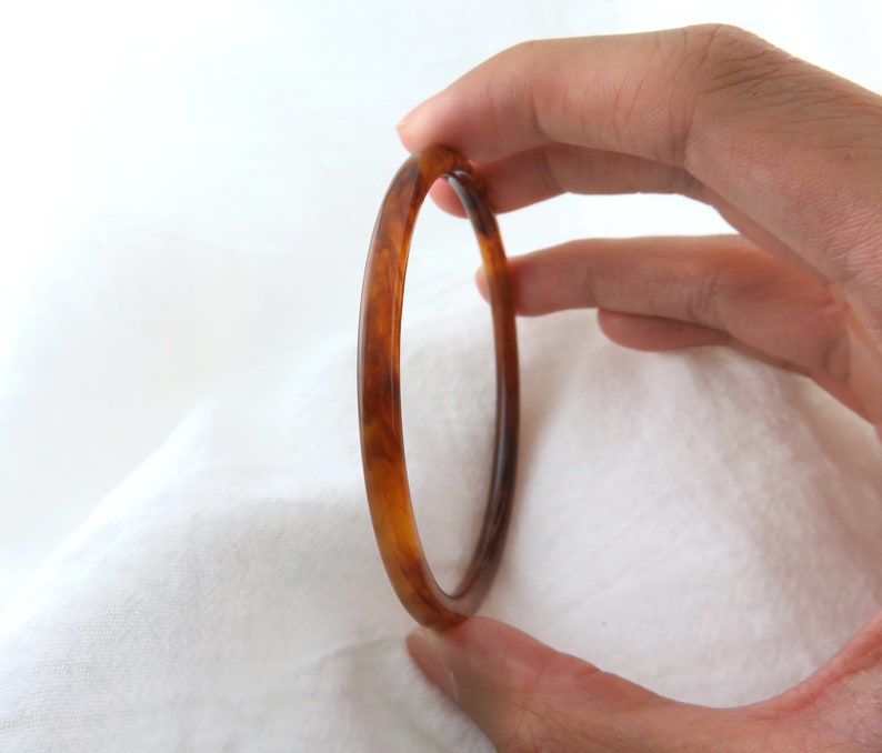 May include: A thin, circular bangle bracelet featuring a polished, translucent amber-colored resin material with a marbled, swirling pattern of dark brown and golden honey tones throughout the smooth, rounded surface of the jewelry piece.
