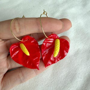 Può includere: Un paio di orecchini rossi a forma di cuore con accenti gialli, che ricordano i fiori di anthurium. Gli orecchini sono sospesi da cerchi dorati. Gli orecchini sono tenuti da una mano.