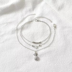 May include: A set of three silver-colored anklets. One anklet features small silver beads, another has a star charm, and the third has a pineapple pendant. The anklets are displayed on a white, textured surface.