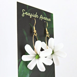 May include: A pair of gold-toned hoop earrings with white flower pendants. The earrings are displayed on a black card with the text "Seaside Avenue" in gold script, against a backdrop of green leaves.