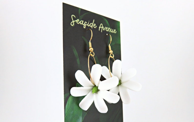 May include: A pair of gold-toned hoop earrings with white flower pendants. The earrings are displayed on a black card with the text "Seaside Avenue" in gold script, against a backdrop of green leaves.
