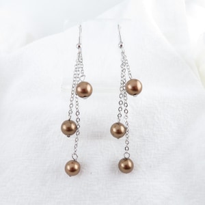 May include: A pair of silver dangle earrings with brown pearl accents. The earrings have three pearls on each side, hanging from a chain.