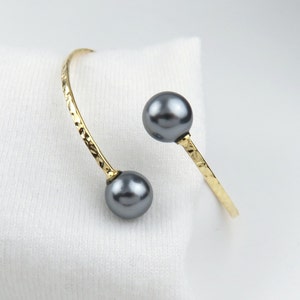 May include: A gold bangle bracelet with two dark gray pearls. The bracelet is textured and has a hammered finish.