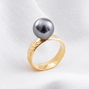 以下が含まれることがあります： 黒真珠が乗った金の指輪。指輪には細かい花柄のデザインが施されています。