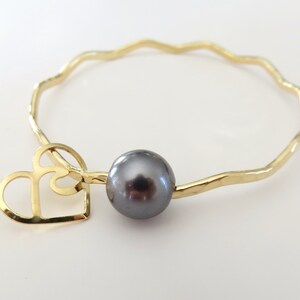 May include: A gold bangle bracelet with a wavy design and a black pearl charm. The charm is attached to a gold heart cutout.