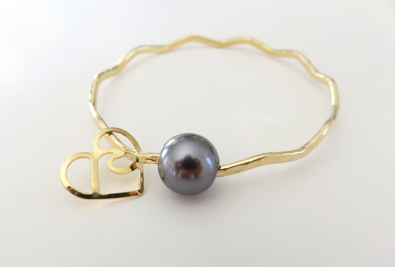 May include: A gold bangle bracelet with a wavy design and a black pearl charm. The charm is attached to a gold heart cutout.