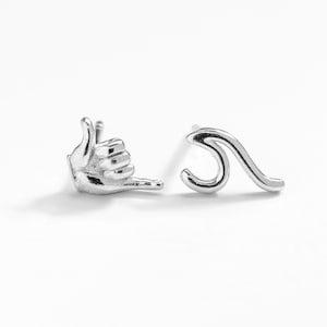 May include: A pair of silver earrings, one shaped like a wave and the other shaped like a hand giving the shaka sign.