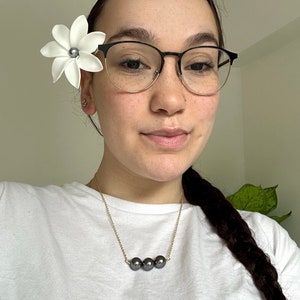May include: A woman wearing a white t-shirt and black framed glasses with a white flower in her hair. She is wearing a gold chain necklace with three black pearls.