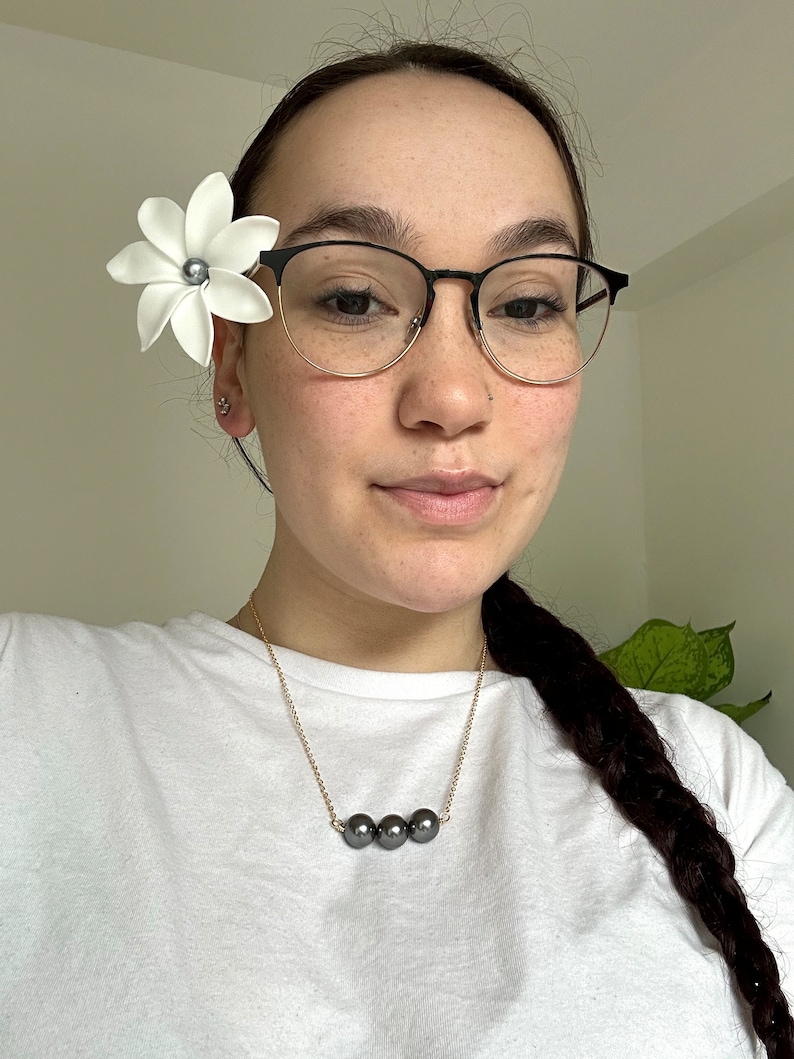 May include: A woman wearing a white t-shirt and black framed glasses with a white flower in her hair. She is wearing a gold chain necklace with three black pearls.