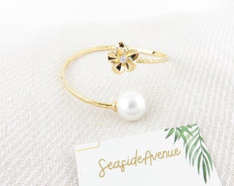 May include: Gold hammered cuff bracelet with a pearl and a flower charm with a rhinestone center. The bracelet is on a white background with a business card that says "Seaside Avenue".