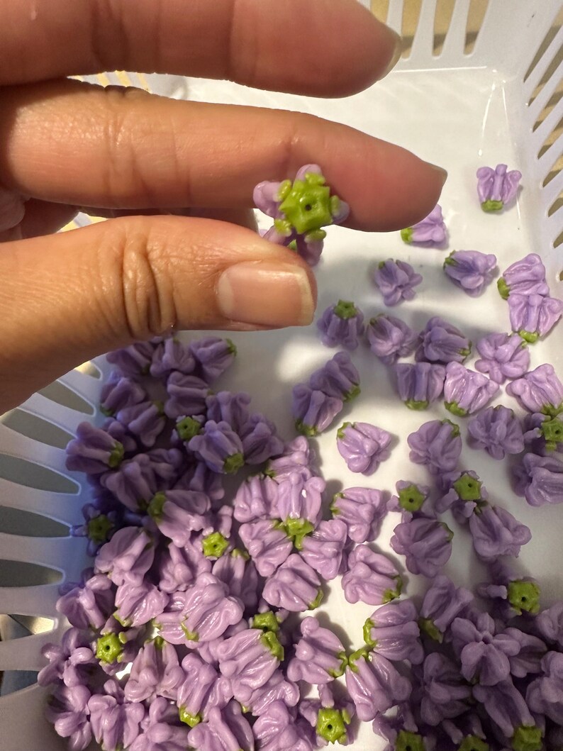 May include: A hand holding a small, lavender-colored flower with a green center. The flower is one of many in a white basket. The flowers have a detailed, textured appearance, suggesting they are handmade or crafted.