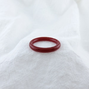 May include: A simple red ring made of smooth, polished material. The ring is lying on a white fabric surface.