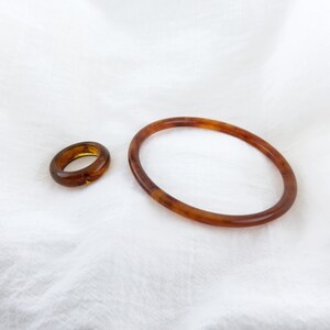 May include: A smooth, polished ring and a matching thin bangle bracelet, both featuring a translucent, warm amber-brown color with subtle, swirling patterns throughout the material, displayed together as a coordinated set of jewelry accessories.