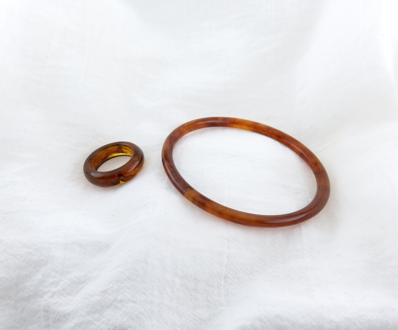 May include: A smooth, polished ring and a matching thin bangle bracelet, both featuring a translucent, warm amber-brown color with subtle, swirling patterns throughout the material, displayed together as a coordinated set of jewelry accessories.