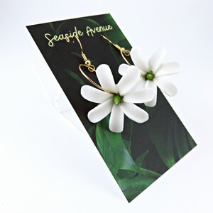 May include: White flower earrings with gold-colored hooks and green centers. The earrings are displayed on a black card with the text "Seaside Avenue" in gold lettering and a green leaf design.