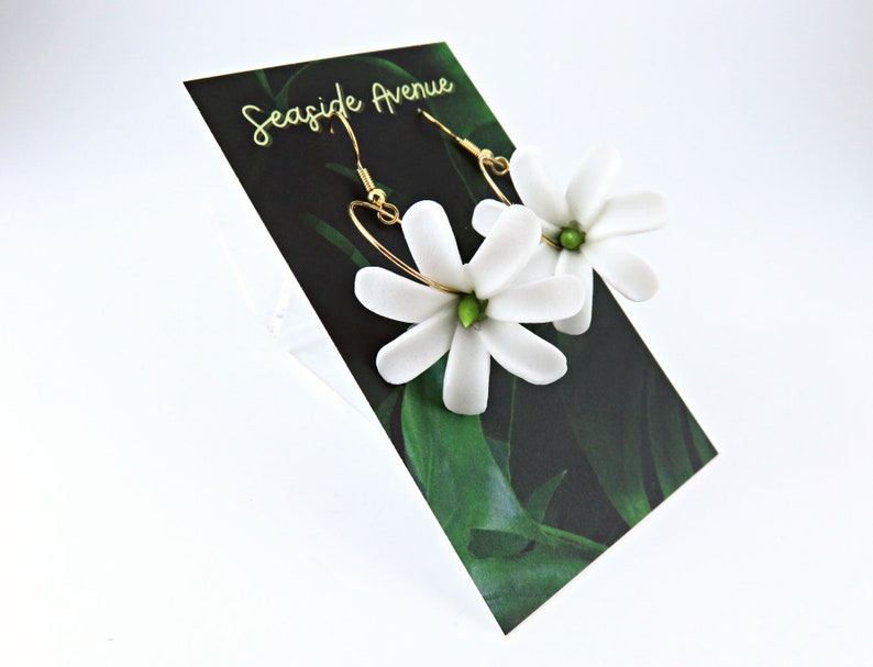 May include: White flower earrings with gold-colored hooks and green centers. The earrings are displayed on a black card with the text "Seaside Avenue" in gold lettering and a green leaf design.