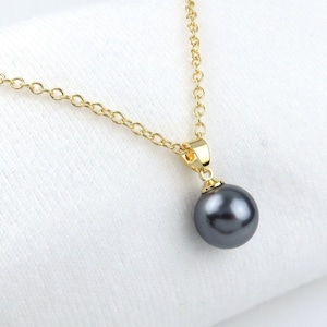 May include: A gold-colored chain necklace with a pendant featuring a dark gray pearl. The pearl is round and hangs from a gold-colored bail. The necklace is displayed on a white surface.