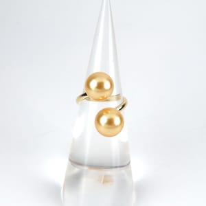 May include: A gold ring with two large, round, golden pearls. The ring is displayed on a clear cone-shaped stand.