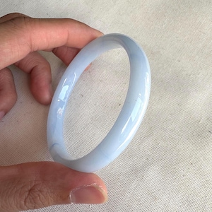 Puede incluir: Un brazalete de jade azul claro. La superficie lisa y pulida refleja la luz, mostrando la calidad translúcida de la piedra. El brazalete tiene forma circular, diseñado para ser usado en la muñeca.