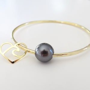 May include: Gold bangle bracelet with a black pearl and a heart charm. The heart charm has a wave design inside.