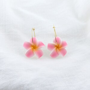 Puede incluir: Un par de pendientes de aro dorados con dijes de flores rosas. Las flores tienen un centro blanco y cinco p&eacute;talos.