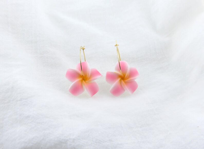 Puede incluir: Un par de pendientes de aro dorados con dijes de flores rosas. Las flores tienen un centro blanco y cinco p&eacute;talos.