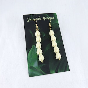 May include: A pair of white flower-shaped dangle earrings with gold hooks. The earrings are displayed on a card with the text "Seaside Avenue".