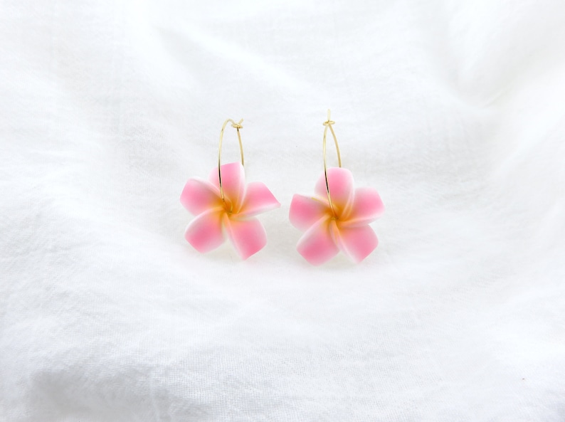 Puede incluir: Un par de pendientes de aro dorados con dijes de flores rosas. Las flores tienen un centro blanco y est&aacute;n hechas de pl&aacute;stico.