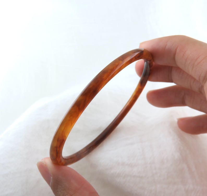 May include: A thin, circular bangle bracelet featuring a translucent, polished finish with a marbled pattern in shades of amber, honey, and dark brown. The bracelet has a smooth, rounded profile and a glossy surface that catches the light.