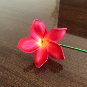 May include: A single pink plumeria flower with a green stem. The flower is on a dark brown wooden surface.