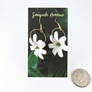 May include: A pair of gold hoop earrings with white flower pendants. The earrings are displayed on a card with the text "Seaside Avenue" and a green leaf design. A quarter is in the foreground.