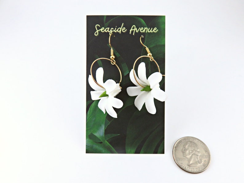 May include: A pair of gold hoop earrings with white flower pendants. The earrings are displayed on a card with the text "Seaside Avenue" and a green leaf design. A quarter is in the foreground.