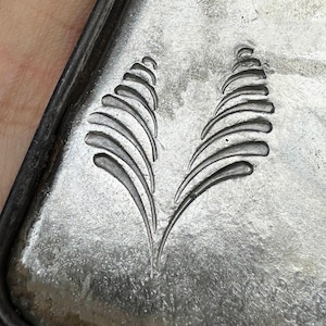 May include: A rectangular silver-colored metal bar featuring a stamped design of two symmetrical, curved, fern-like leaf patterns. The surface of the metal has a brushed, textured finish with a dark, oxidized border along the outer edge.