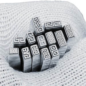Puede incluir: Un juego de 12 sellos metálicos con un diseño de llave griega. Los sellos están dispuestos en una pila y se encuentran en una bolsa de tela blanca.