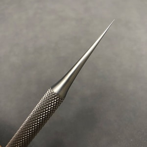 K&ouml;nnte beinhalten: Nahaufnahme eines silberfarbenen Metallahls mit einer scharfen Spitze. Der Griff hat eine strukturierte, rautenf&ouml;rmige Oberfl&auml;che. Das Werkzeug ist f&uuml;r Pr&auml;zisionsarbeiten konzipiert.