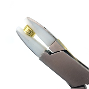 May include: Close-up of a silver and gold jewelry pliers with white plastic jaws holding a gold jump ring.