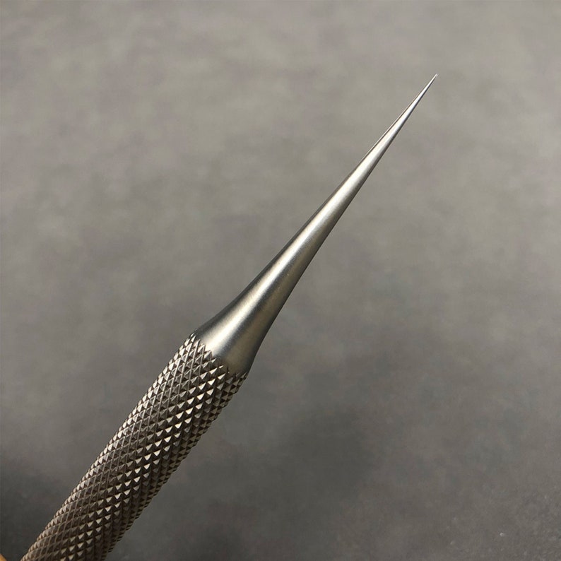 K&ouml;nnte beinhalten: Nahaufnahme eines silberfarbenen Metallahls mit einer scharfen Spitze. Der Griff hat eine strukturierte, rautenf&ouml;rmige Oberfl&auml;che. Das Werkzeug ist f&uuml;r Pr&auml;zisionsarbeiten konzipiert.