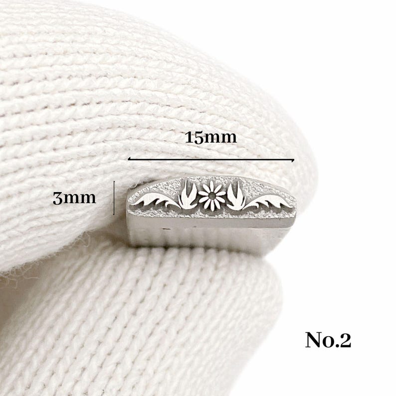 Op de afbeelding: Een metalen stempel met een bloemmotief, 15 mm lang en 3 mm hoog. De stempel heeft een centrale bloem geflankeerd door gestileerde bladeren. Het nummer "No.2" is rechtsonder zichtbaar.