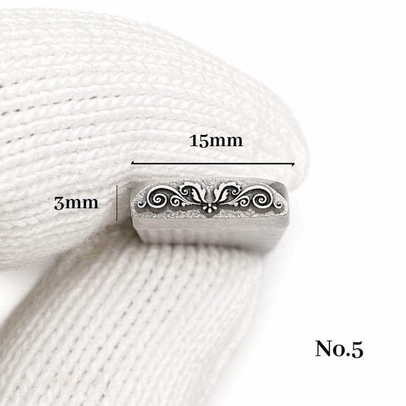 Op de afbeelding: Een rechthoekige metalen stempel met een sierlijk bloemmotief, 1,5 cm lang en 3 mm breed. De stempel is gemarkeerd met "No.5" en wordt tegen een witte stoffen achtergrond gehouden.