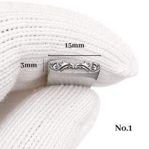 Op de afbeelding: Een zilveren metalen stempel met een sierlijk bloemmotief, 15 mm lang en 3 mm breed. De stempel wordt vastgehouden door een gehandschoende hand. De tekst "No.1" is zichtbaar.