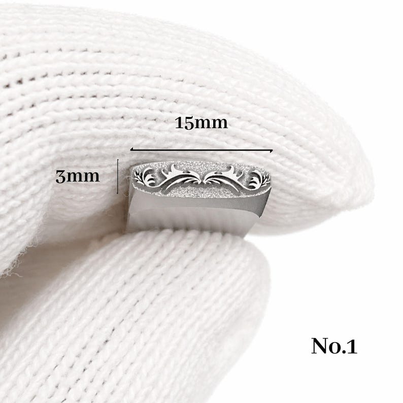 Op de afbeelding: Een zilveren metalen stempel met een sierlijk bloemmotief, 15 mm lang en 3 mm breed. De stempel wordt vastgehouden door een gehandschoende hand. De tekst "No.1" is zichtbaar.