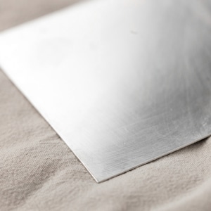 May include: A smooth, silver-colored metal sheet with a brushed finish. The sheet is lying on a beige fabric surface.