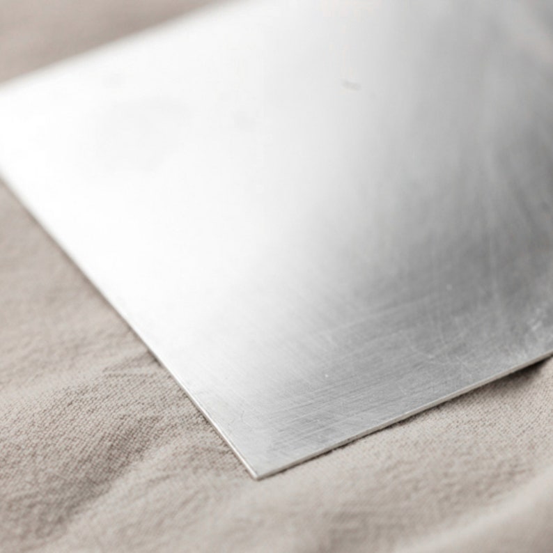May include: A smooth, silver-colored metal sheet with a brushed finish. The sheet is lying on a beige fabric surface.