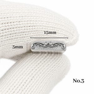 Op de afbeelding: Een klein, rechthoekig zilverkleurig metalen object met een sierlijk, symmetrisch ontwerp. Het object is 1,5 cm lang en 0,3 cm breed. Het nummer "No.3" staat rechtsonder gedrukt. Het object wordt vastgehouden door een gehandschoende hand.