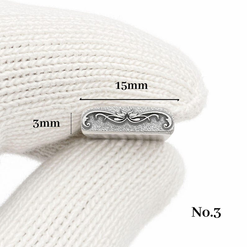 Op de afbeelding: Een klein, rechthoekig zilverkleurig metalen object met een sierlijk, symmetrisch ontwerp. Het object is 1,5 cm lang en 0,3 cm breed. Het nummer "No.3" staat rechtsonder gedrukt. Het object wordt vastgehouden door een gehandschoende hand.