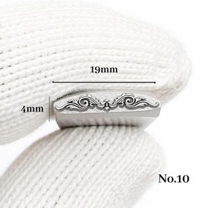 Op de afbeelding: Een metalen stempel met een sierlijk, symmetrisch ontwerp, 1,9 cm lang en 0,4 cm breed. De stempel heeft een decoratief krulpatroon. Het nummer "No.10" staat rechtsonder afgedrukt.